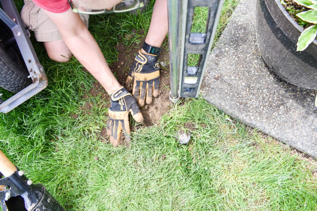 DIY string light pole being installed in the ground, hands packing soil in around the base of the PVC pipe in the ground