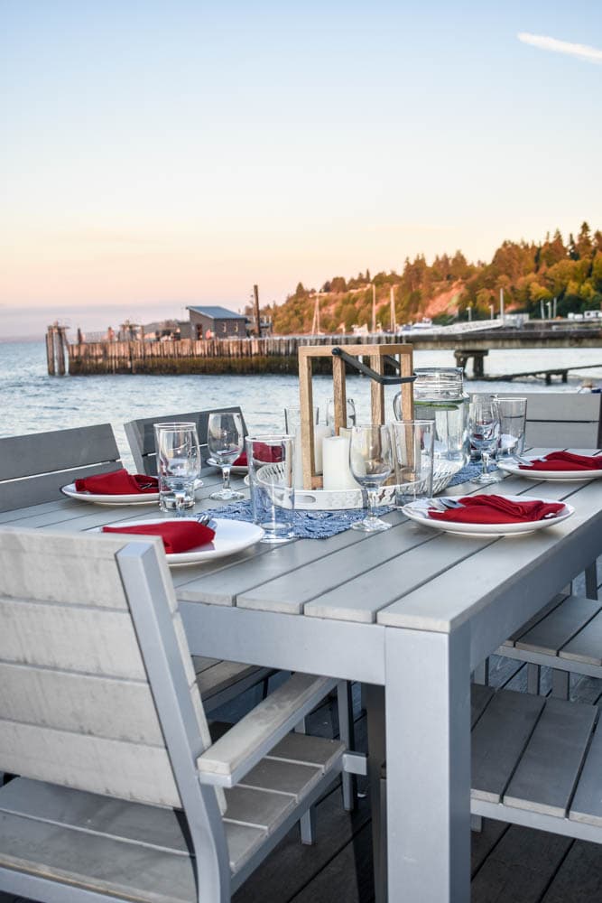 outdoor dining table set for a 4th of July party with a navy blue and white patterned table runner, red cloth napkins, candle lanterns, and a pier in the background