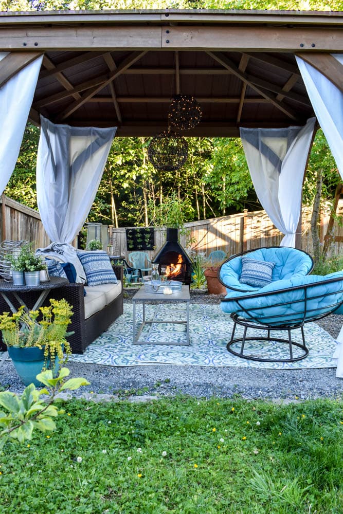 This covered outdoor living room on a budget is the perfect backyard Summer spot to relax! It's got a fireplace to roast marshmallows, cozy modern furniture, and an outdoor rug.