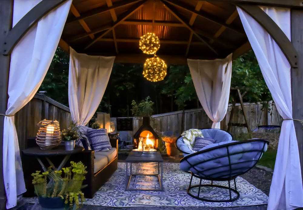 nighttime photo of 12'x12' Costco gazebo by Yardistry Structures as an outdoor living room, with outdoor curtains, papasan chairs, an outdoor sofa, and chandelier