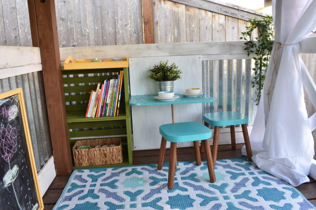image of inside of playhouse with mini dining table and flisat stools, bookshelf, and chalkboard