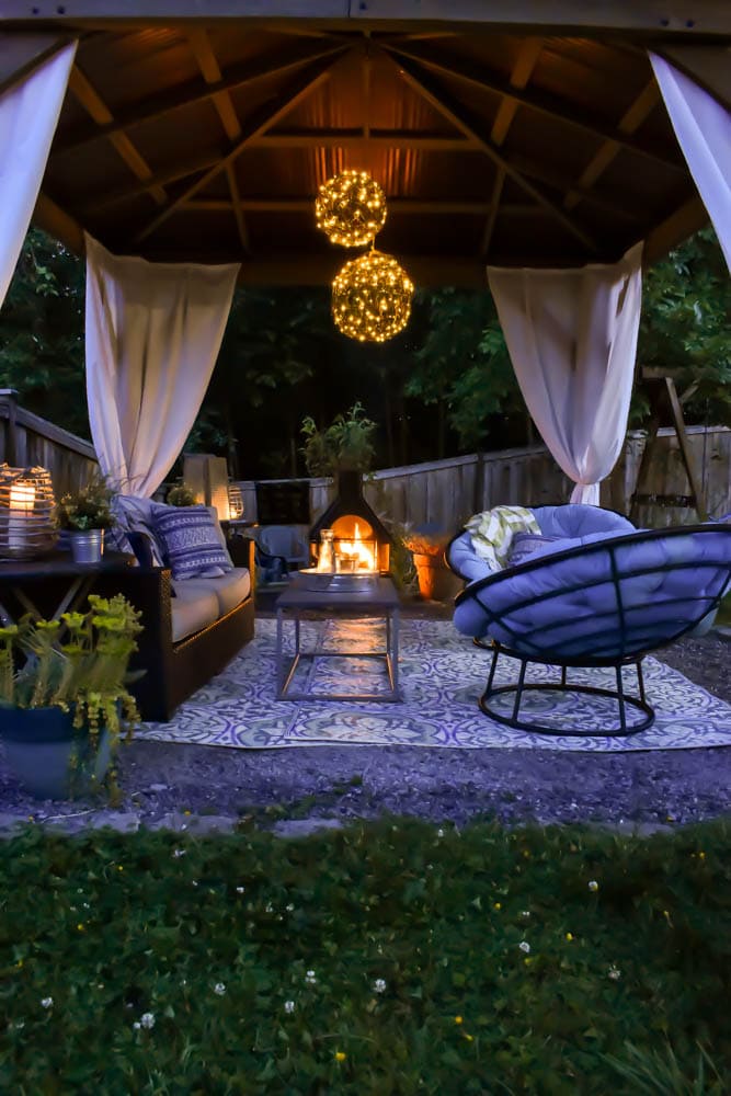 This covered outdoor living room on a budget is the perfect backyard Summer spot to relax! It's got a fireplace to roast marshmallows, cozy modern furniture, and an outdoor rug.