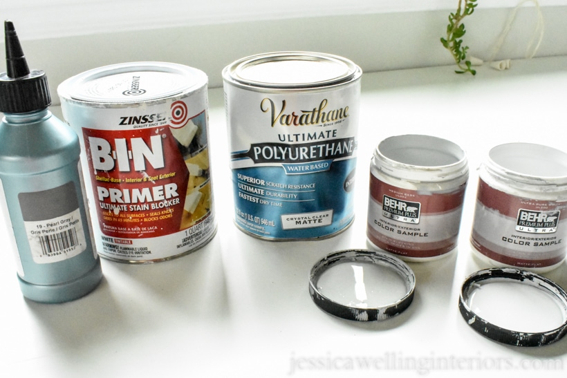 Five paint cans and samples lined up for DIY tile painting project- dark grey grout refresh, BIN primer, polyurethane clear coat, and two Behr paint samples in white and grey