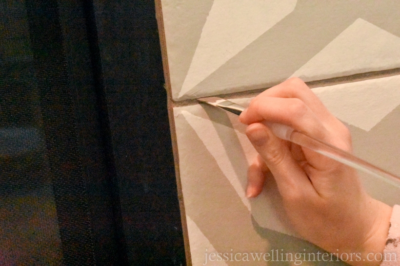 hand holding small angled artist's brush, touching up dark grey grout lines between faux painted cement tiles