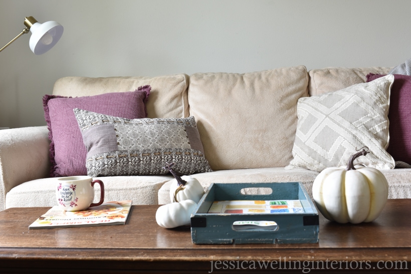 neutral and plum purple Fall living room decor with cream colored sofa, plum and white pillows, and white pumpkins on wood coffee table in foreground