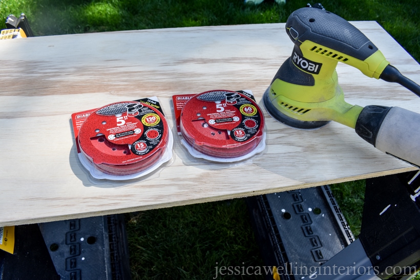 cut plywood piece sitting on two sawhorses, with orbital sander and sandpaper discs sitting on top of it