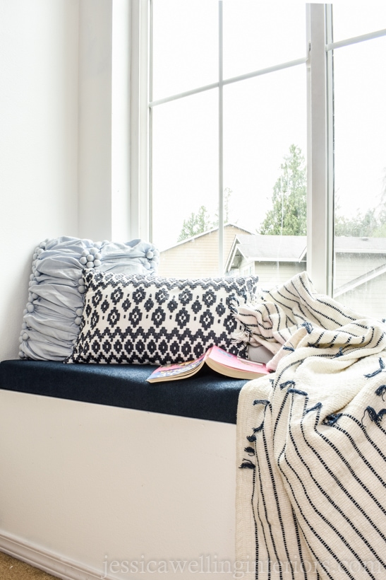 narrow window seat in children's bedroom with upholstered bench cushion, throw pillows, a book, and a blanket