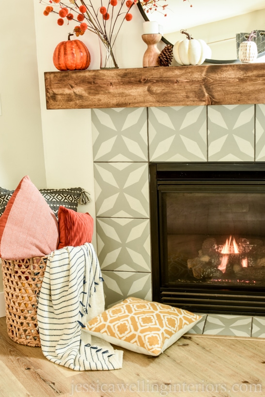 side-view of Fall mantle decor with stems, pumpkins, pine cones, and a basket of Fall throw pillows & blankets on the floor