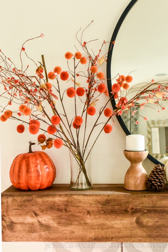 orange and red pom-pom stems in a glass vase sitting on a wood mantle decorated for Fall with a round mirror, candle holder, and orange pumpkins