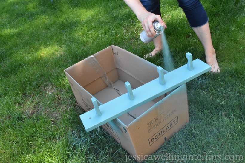 woman applying second coat of aqua-colored spray paint to modern coat hooks rack outdoors in grass