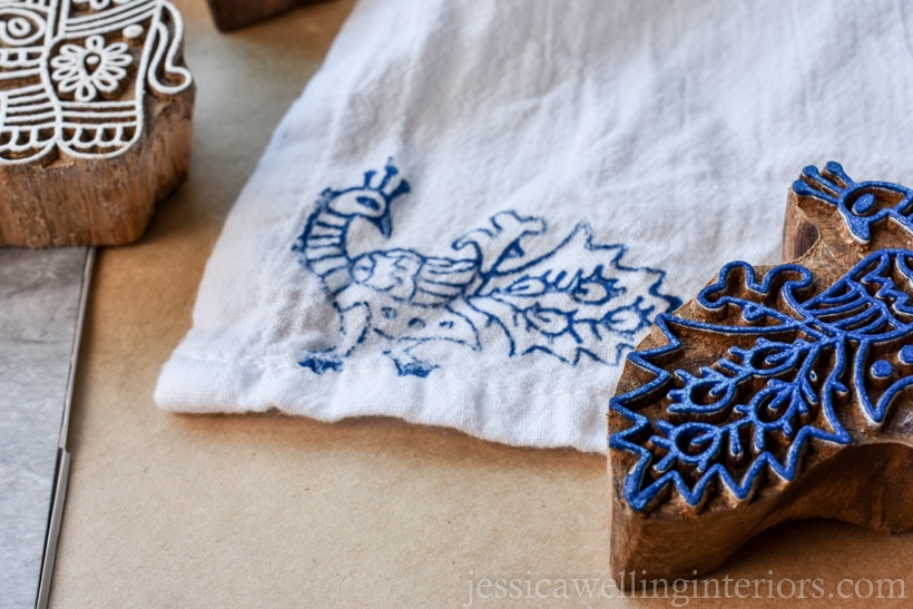 close-up of handmade wood printing block peacock with blue paint on it, and tea towel stamped with the same in the background