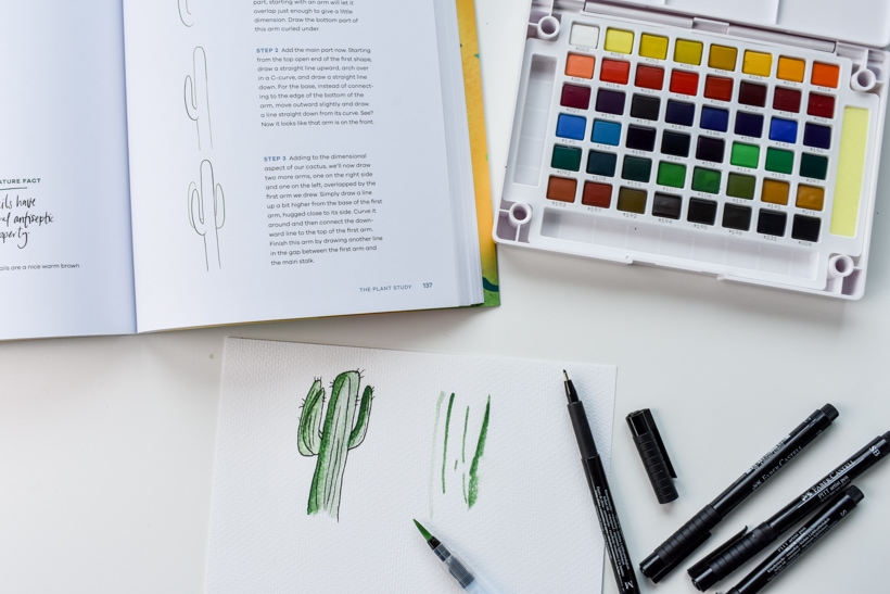 watercolor paint palette next to open workbook and paper with watercolor cactus and black drawing pens gift ideas for women