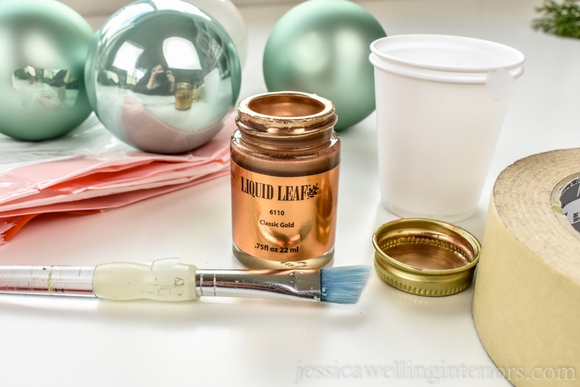 supplies to make DIY Christmas ornaments- glass ball ornaments, gold leaf paint, paint brush, plastic trash bag, masking tape, and dixie cups