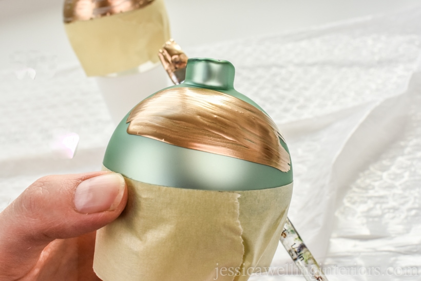 hand holding glass ball ornament wrapped with masking tape and painting gold leaf paint on the exposed part of the ornament