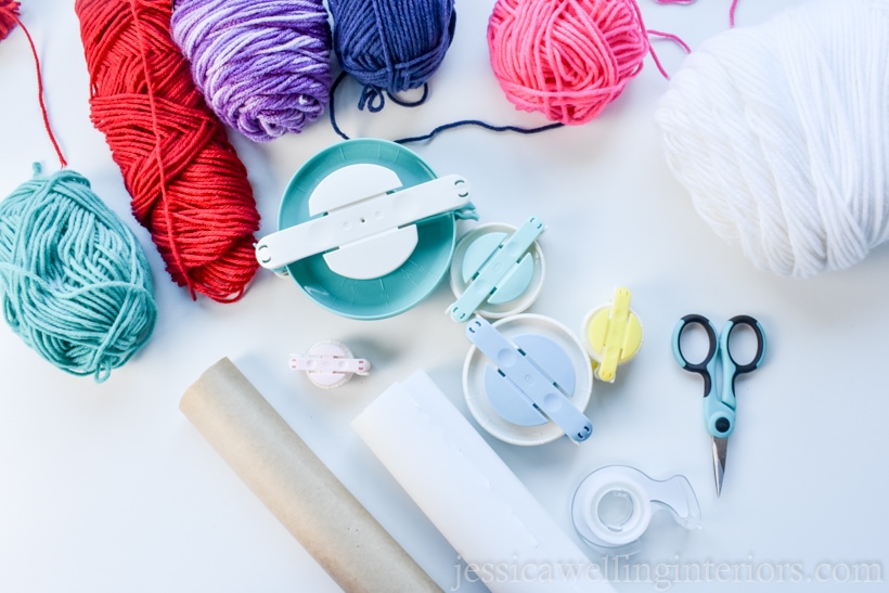 flatlay photo of supplies needed to make pompoms and wrap gifts- colorful arn skeins, pom pom makers in 5 different sizes, tape, craft scissors, and wrapping paper