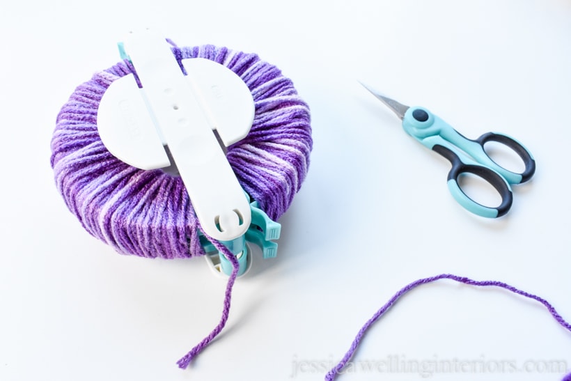 large pom pom maker with purple yarn wrapped around it and a pair of blue craft scissors