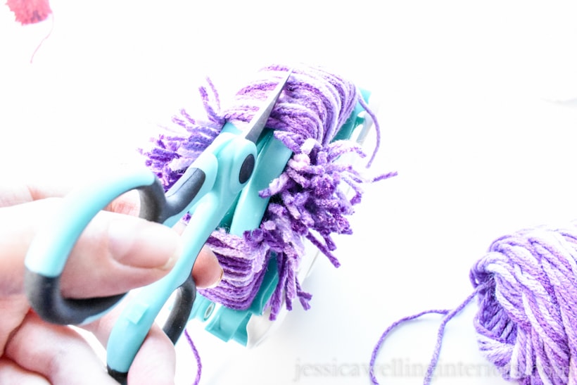 close-up of hand using craft scissors to cut purple yarn on pom pom maker