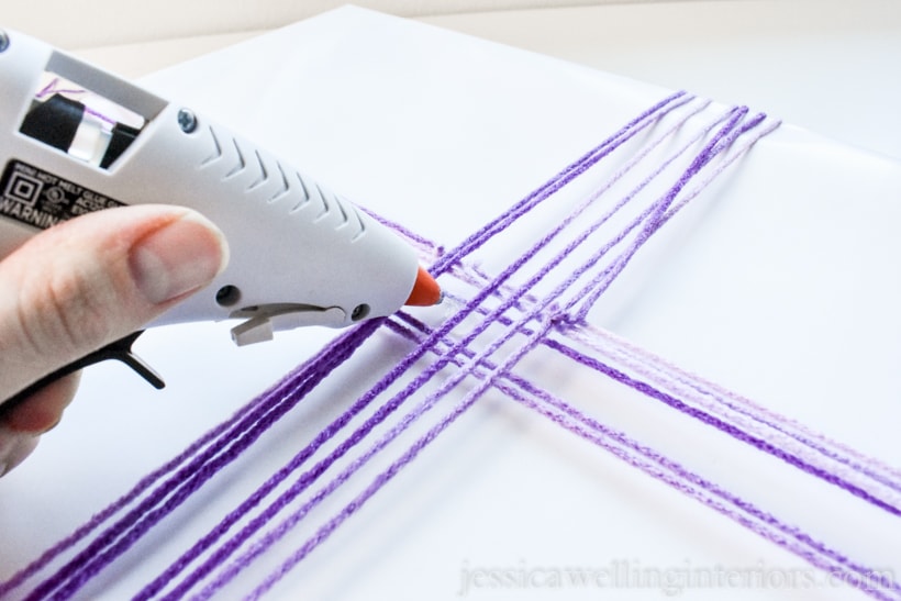 hand holding hot glue gun- applying glue to gift wrapped with white paper and tied with purple yarn