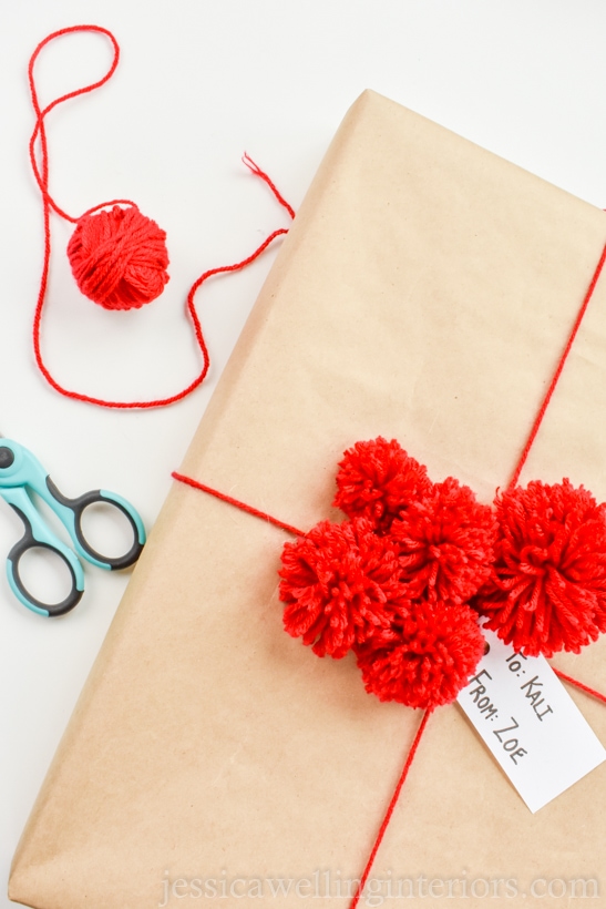 brown paper-wrapped Christmas gift, tied with red yarn and topped with a cluster of several red pom poms of different sizes