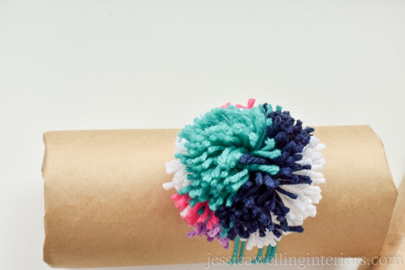 cylider-shaped gift wrapped in brown craft paper and tied with a large multi-coloed pom pom