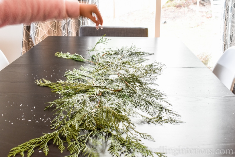 table with cedar branches down the center, and child's hand sprinkling fake snow on them