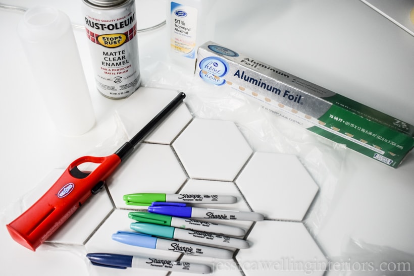 diy coasters tile supplies: hexagon ceramic tiles, aluminum foil, lighter, blue and green Sharpie pens, clear enamel spray paint, isopropyl alcohol, and a squeeze bottle