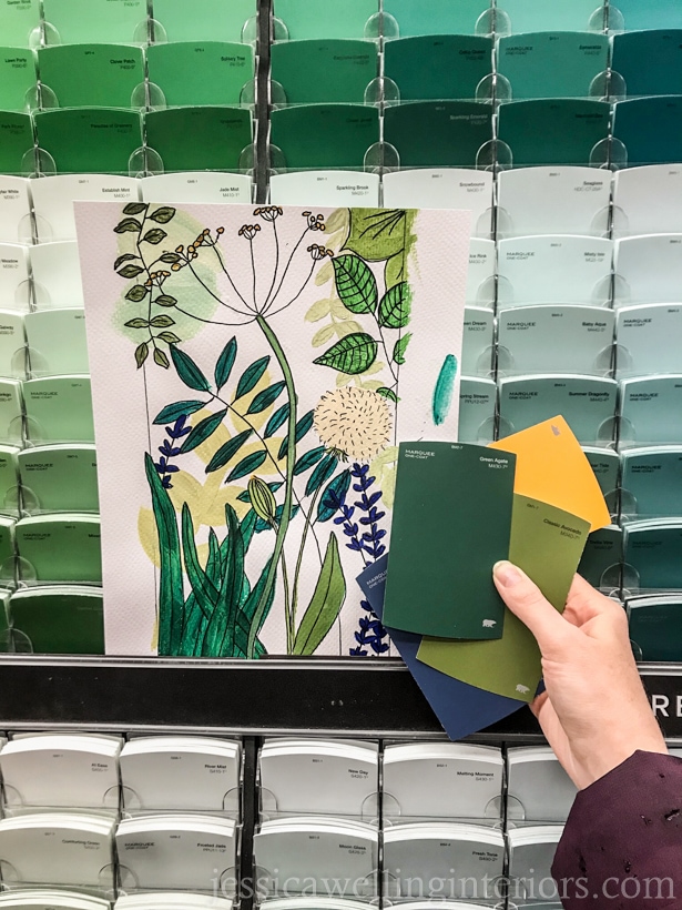 hand drawn and painted botanical art held up in front of paint swatch rack at Home Depot, with hand holding several paint swatchws in greens, yellow, and blue