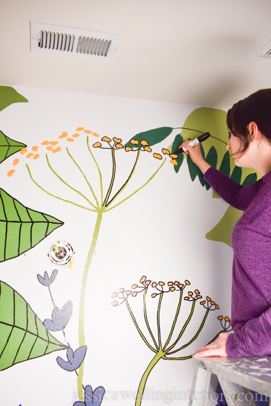 woman standing on ladder, outlining botanical mural painting with a black paint pen