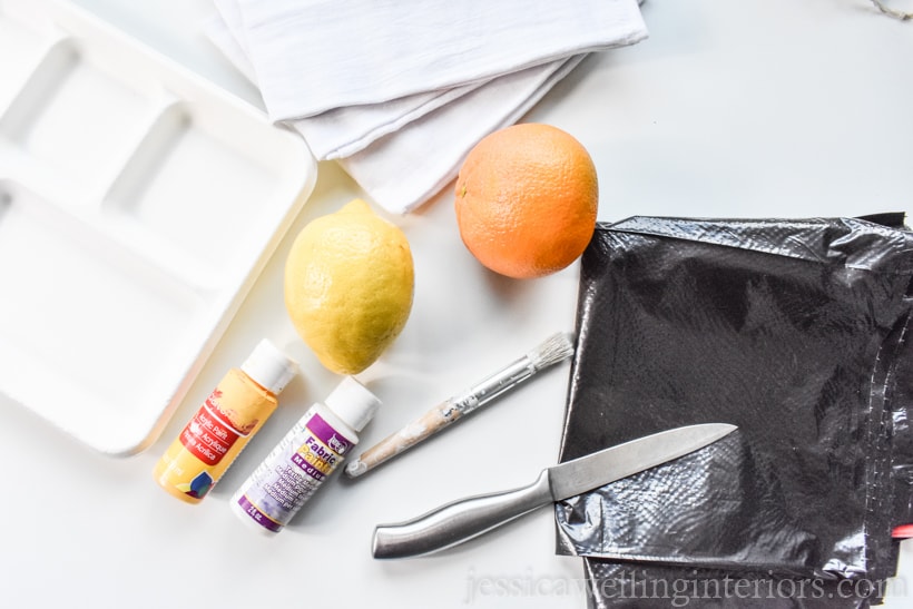 materials to make easy DIY hand-printed tea towels: orange, lemon, plastic trash bag, knife, acrylic paint, paper plate, and white flour sack dish cloths