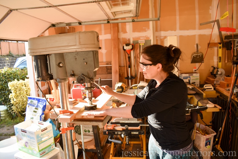 woman using a drill press and hole saw to cut holes in board to hold indoor hanging plants in terra cotta pots