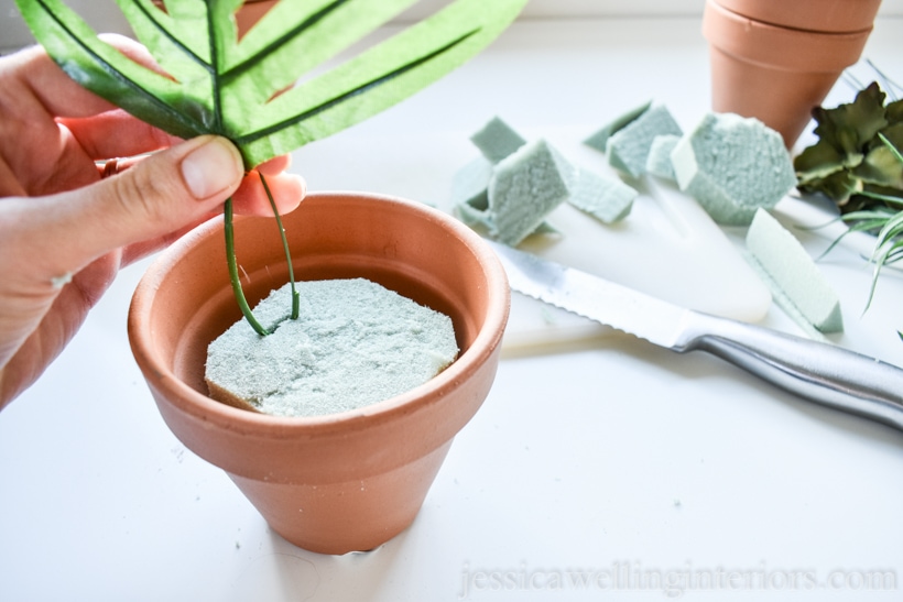 hand placing faux plant stem into floral foam in a terra cotta pot