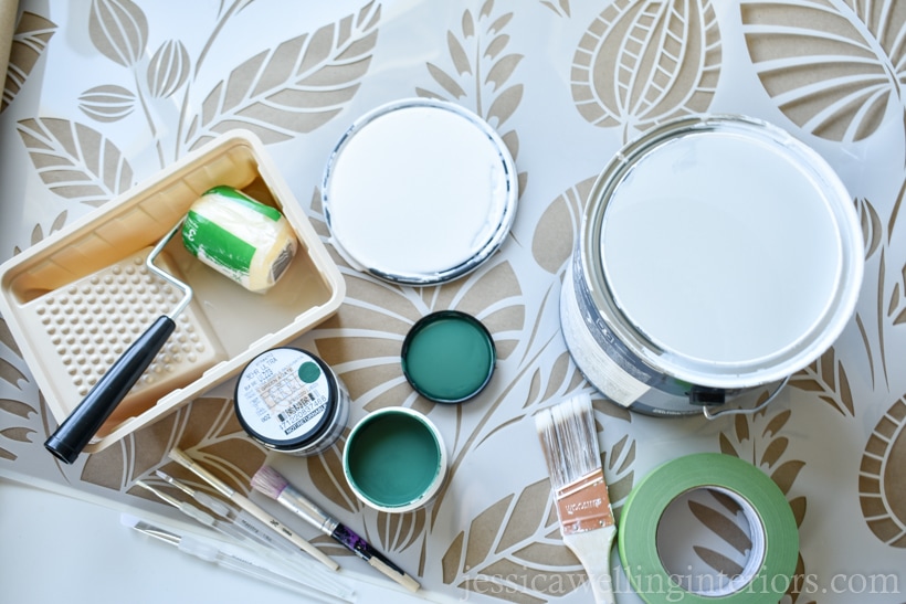 overhead view of materials need to stencil a wall with paint: small paint roller & tray, wall paint, small paintbrushes, a large paintbrush, and Frog tape