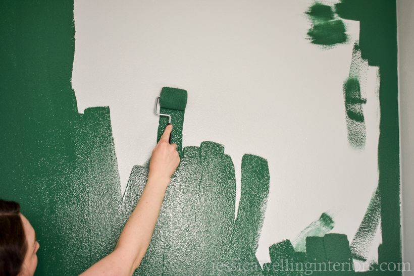 close-up of a woman using a small paint roller to apply dark green paint to an accent wall