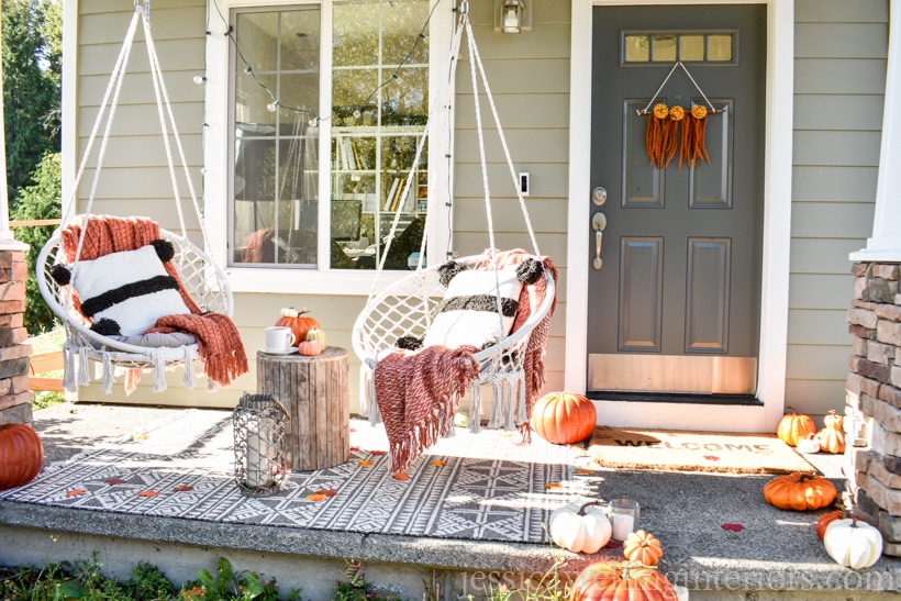 small front porch decorated for Fall- modern macrame swing chairs with cozy throw pillows and blankets, pumpkins, string lights, and a Boho wall hanging on the front door