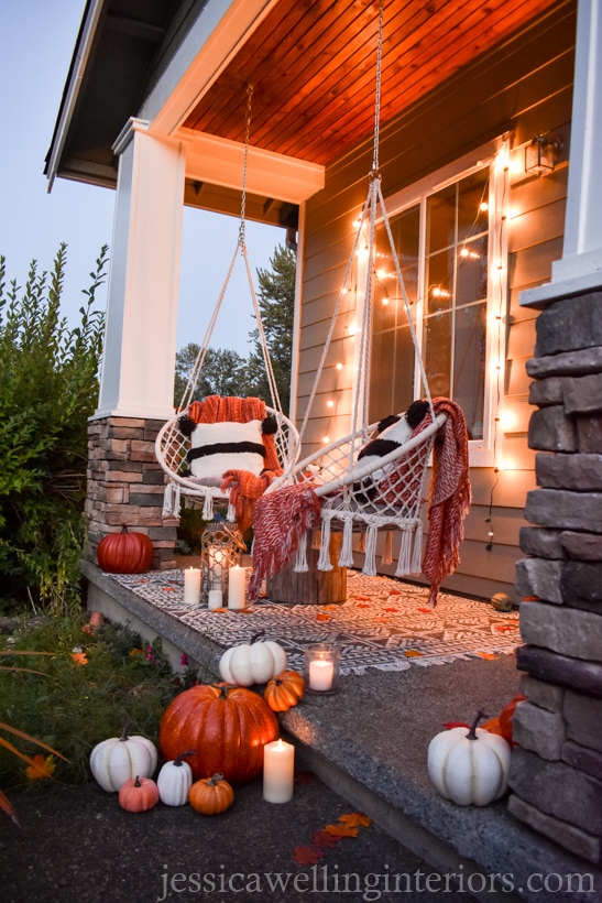 low-light photo of Fall porch decor with modern Boho swings, pumpkins, throw pillows, and blankets, candles, and string lights