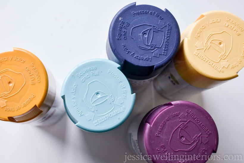 top view of five cans of spray paint in mustard yellow, purple, navy blue, teal, and light yellow