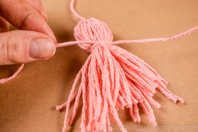 hand tying a pink yarn tassel around the center with another piece of pink yarn