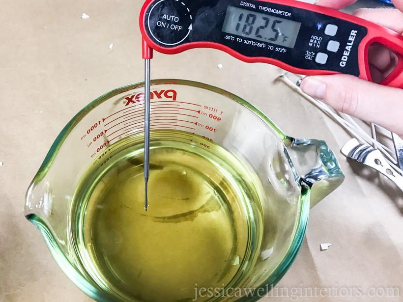 close-up of glass measuring cup filled with melted soy wax and a hand holding a digital thermometer, which reads 182.5 degrees F