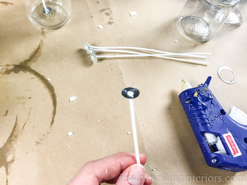 hand holding a cotton candle wick with the metal end in the air and a hot glue gun in the background