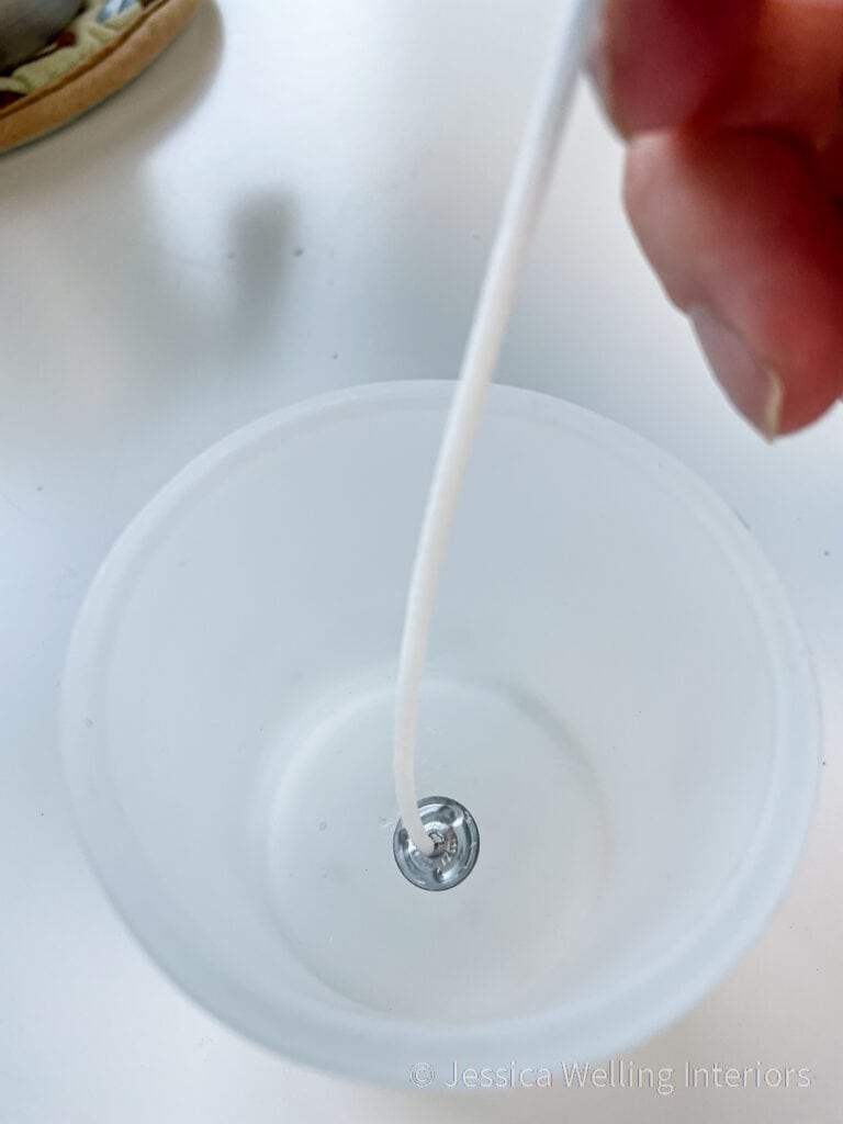 small glass container with a candle wick being attached to the bottom