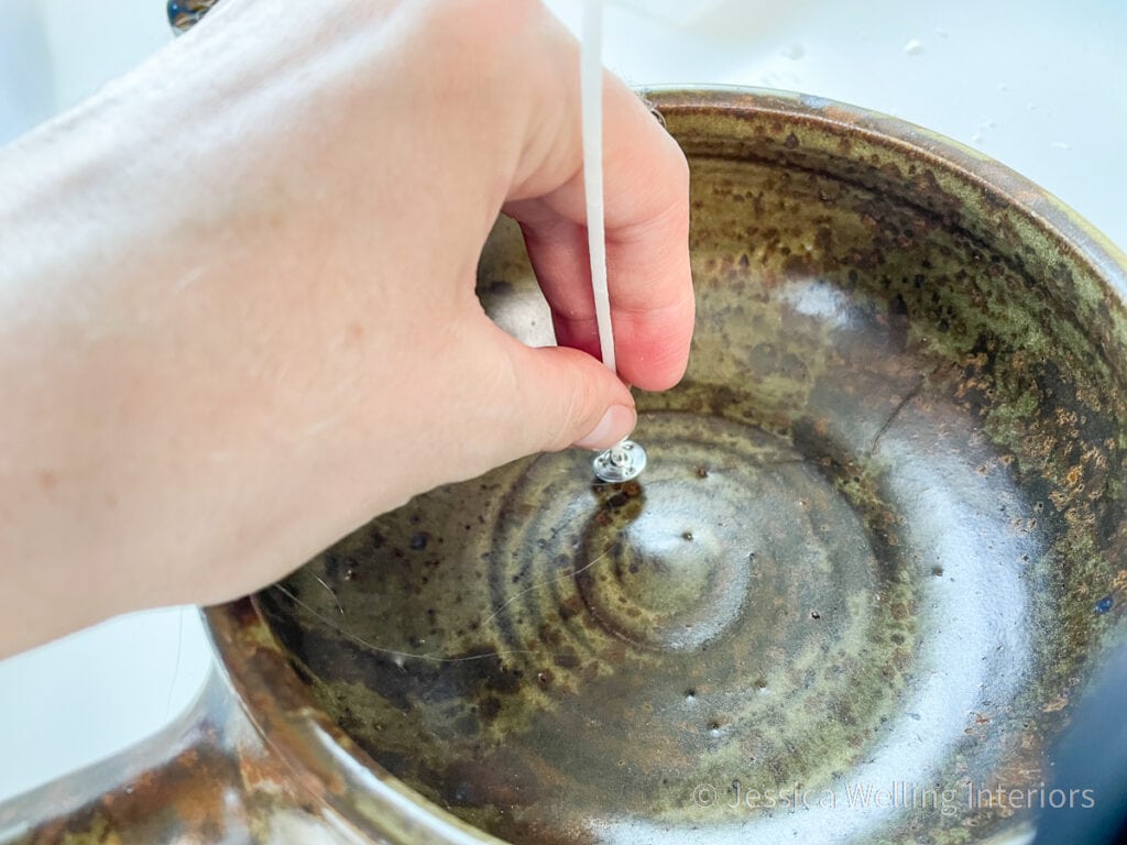 close-up of hand attaching the bottom of a cotton candle wick to a rustic bowl with hot glue