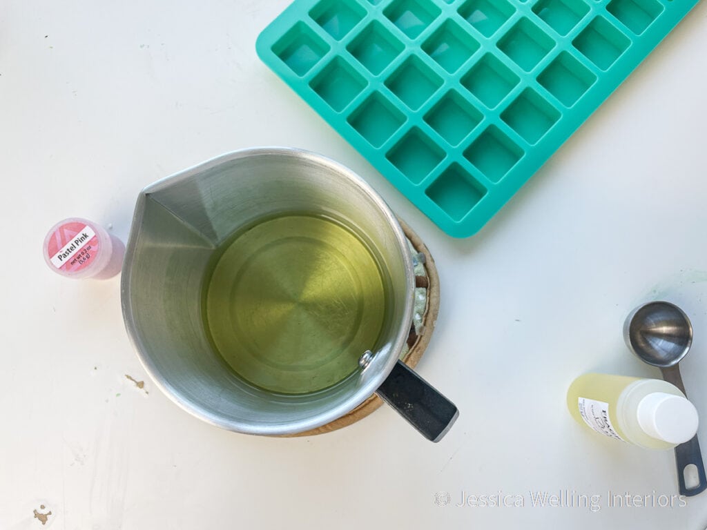 overhead view of metal pitcher of melted soy wax, silicone wax melt mold, fragrance oil, and container of pink wax dye chips