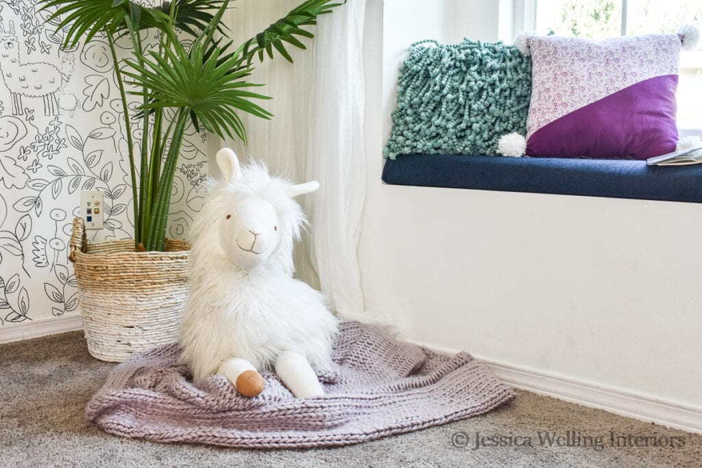 white stuffed llama sitting on the floor of a girls' bedroom with a purple blanket and a tall faux plant