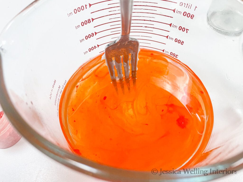 glass measuring cup of melted soy wax to make candles with orange dye being stirred in with a fork