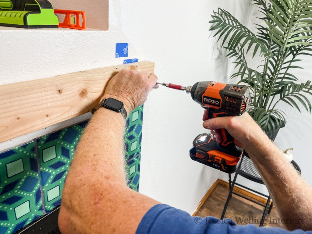 hand using a drill to attach a 2x4 to the wall to mount a DIY fireplace mantel to the wall
