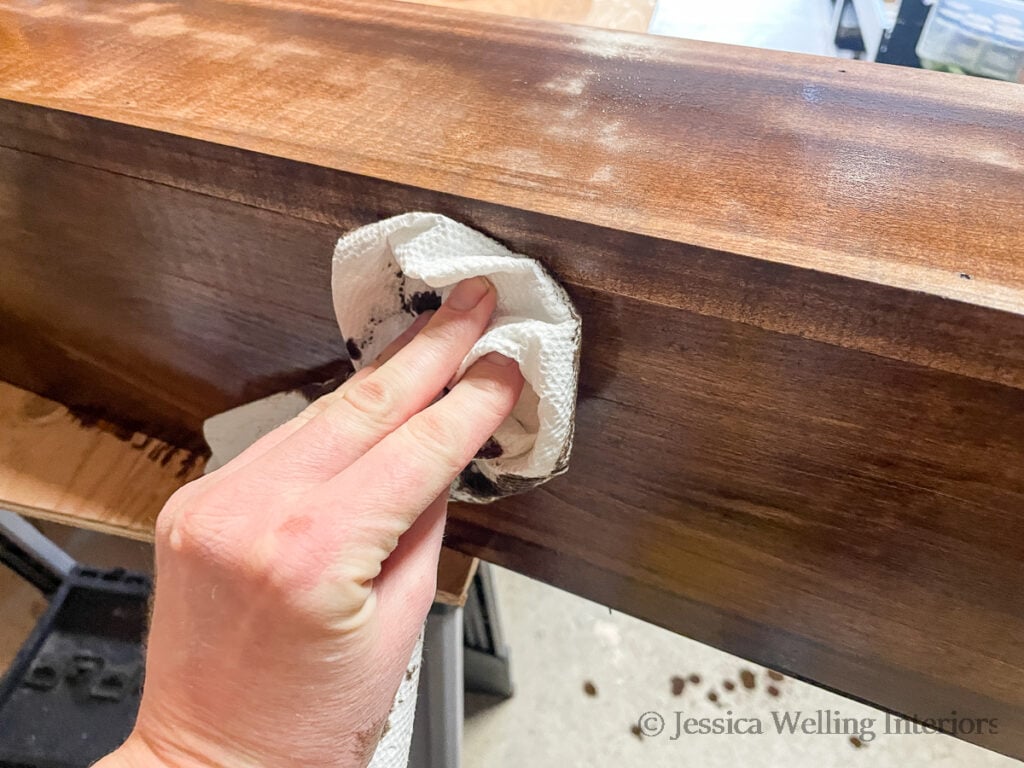 close-up of a hand using a paper towel to wipe wood stain off of a DIY mantel