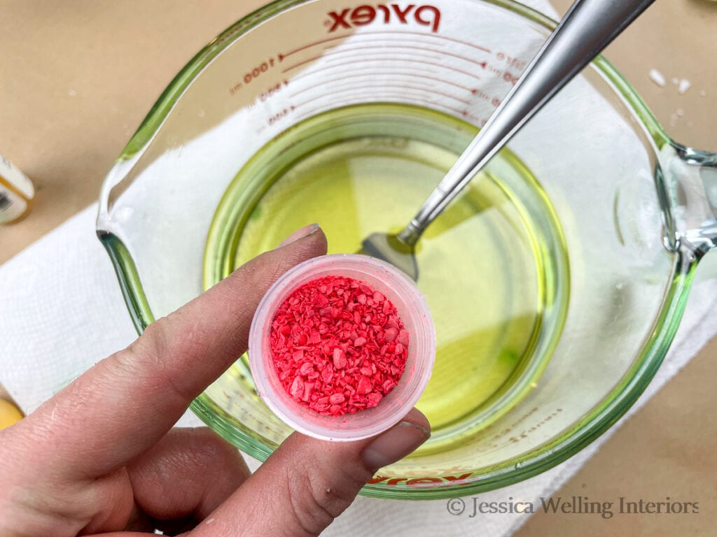 hand holding a container of hot pink wax dye chips above a container of melted soy wax to make DIY wax melts