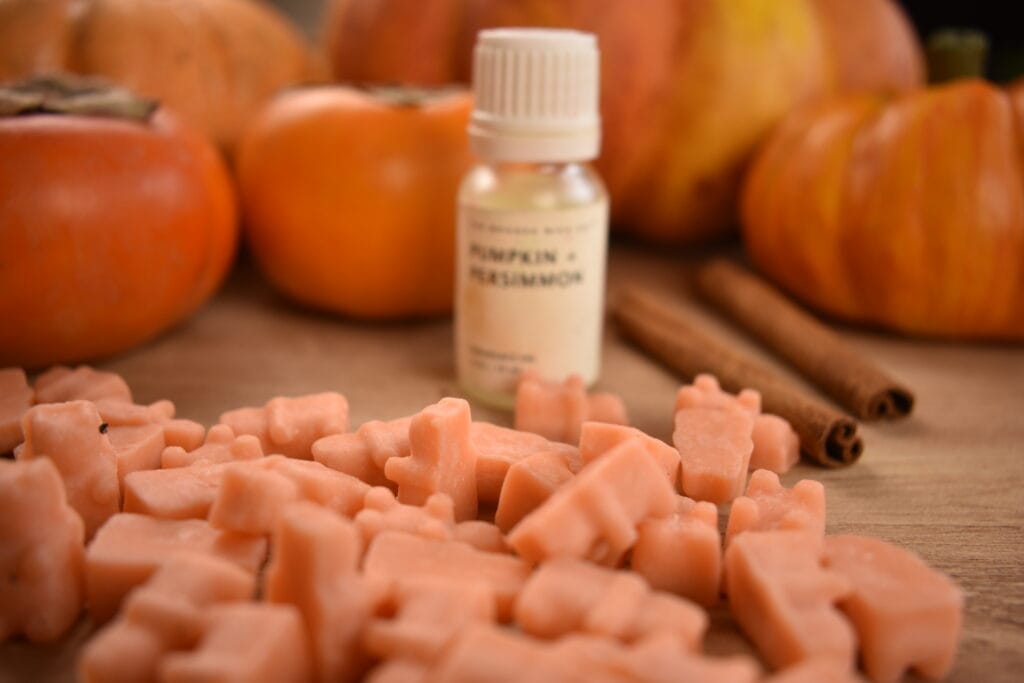 orange gummy bear shaped wax melts with a bottle of Pumpkin & Persimmon candle fragrance oil from The Wooden Wick Co., pumpkins, and cinnamon sticks