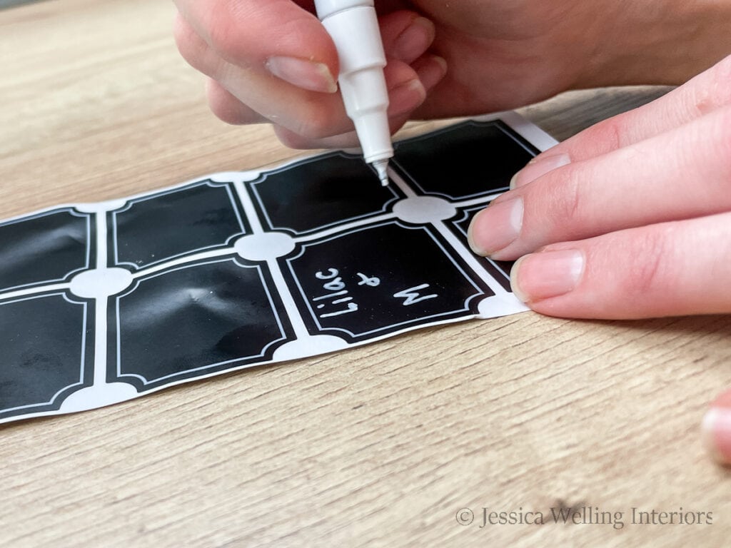 hands using a white paint pen to write "Lilac & Mimosa" on a chalkboard sticker label
