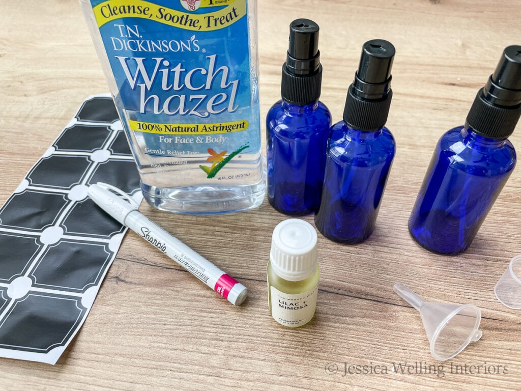 materials for making room spray- blue glass spray bottles, witch hazel, fragrance oil, a small funnel, chalkboard sticker labels, and a white paint pen